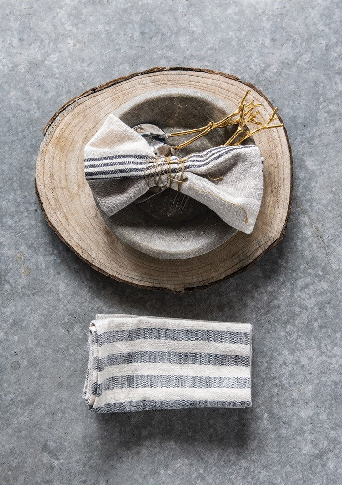 woven cotton striped napkins displayed with a bowl and flatware on a round wood cookie slab on a gray surface