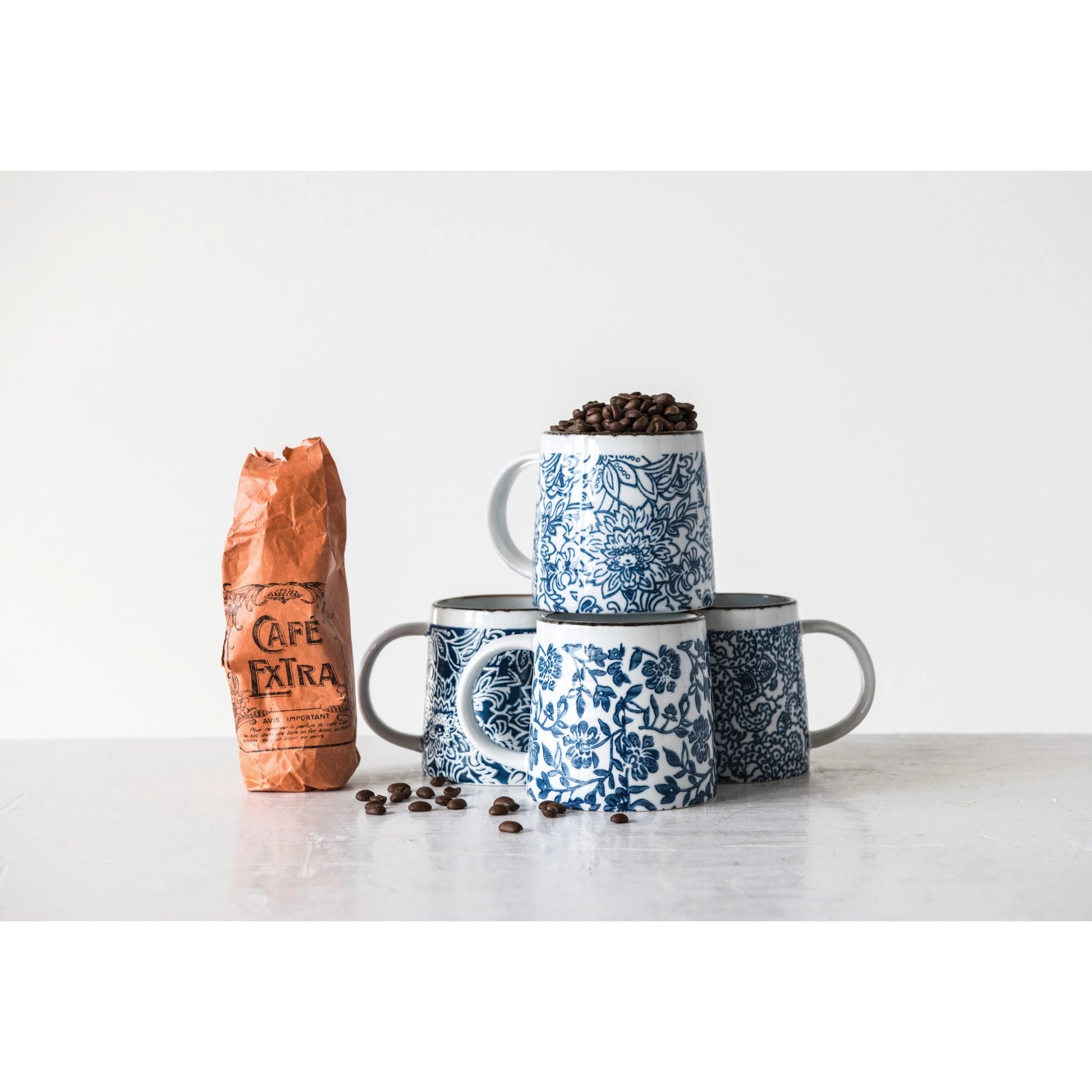 4 blue patterned mug stacked and arranged with a bag of coffee and coffee beans scattered along a table.