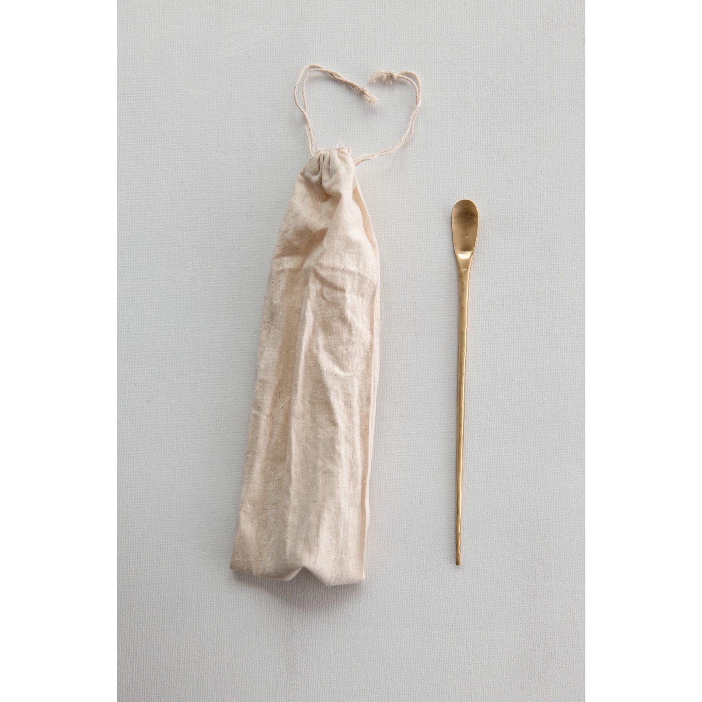 brass cocktail spoon displayed next to the dust bag on a light gray background