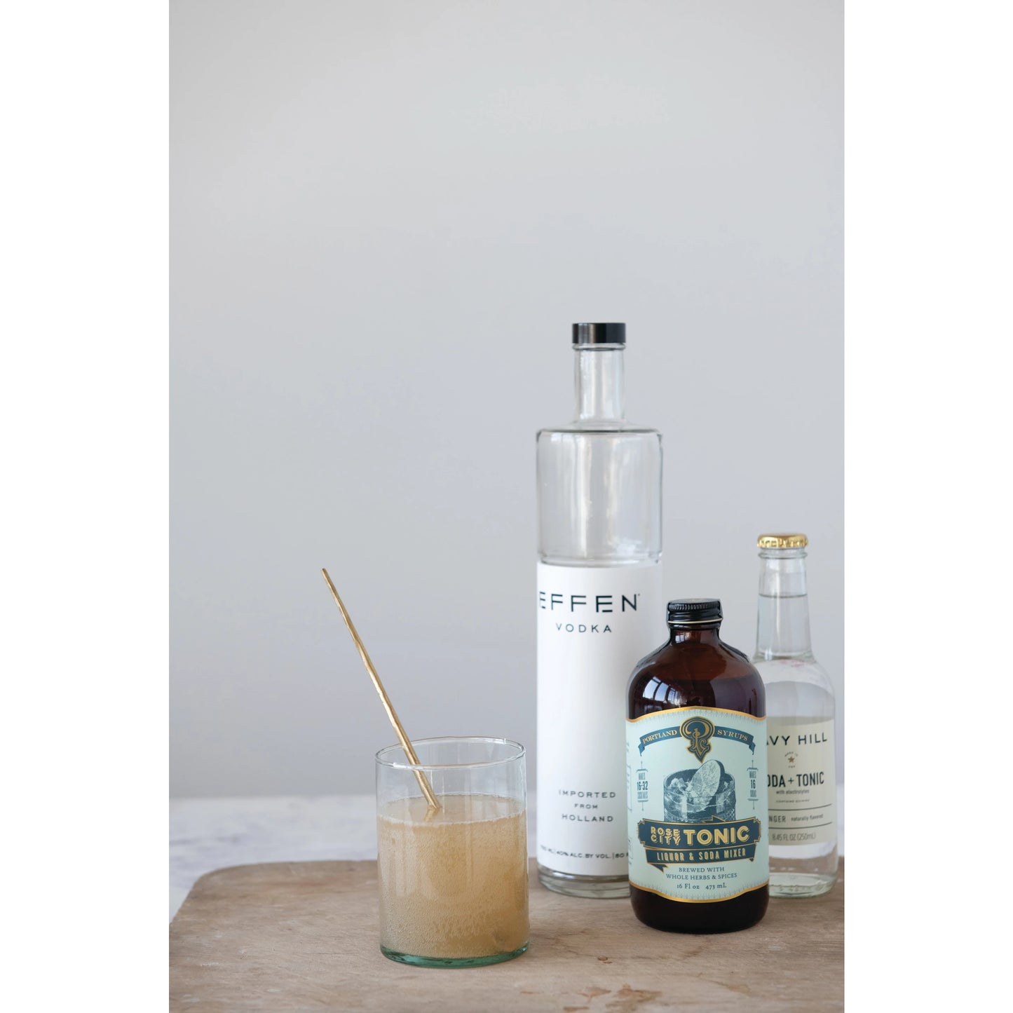 brass cocktail spoon displayed next to bottle of drink mixes on a wood board against a light gray background