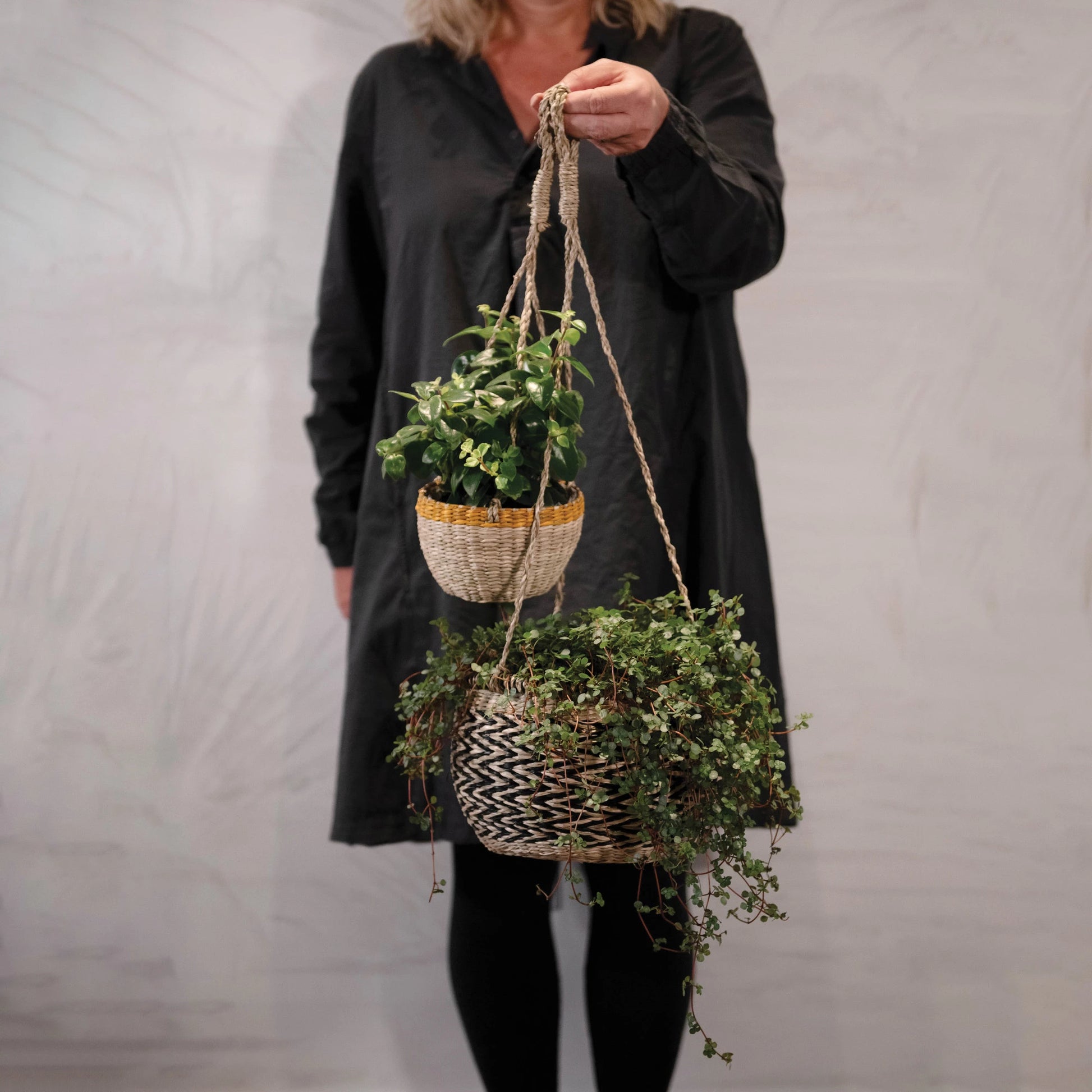 a woman holding a large and small hand woven hanging basket planters filled with plants out in front of her