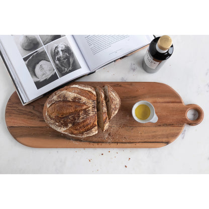 oval acacia wood cutting board with handled displayed with bread, small bowl of dipping oil, and book on a white surface
