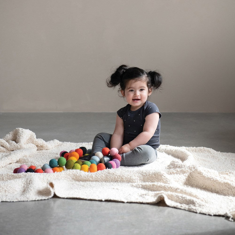 a little girl sitting on a white blanket playing with the wool felt rainbow garland against a gray background