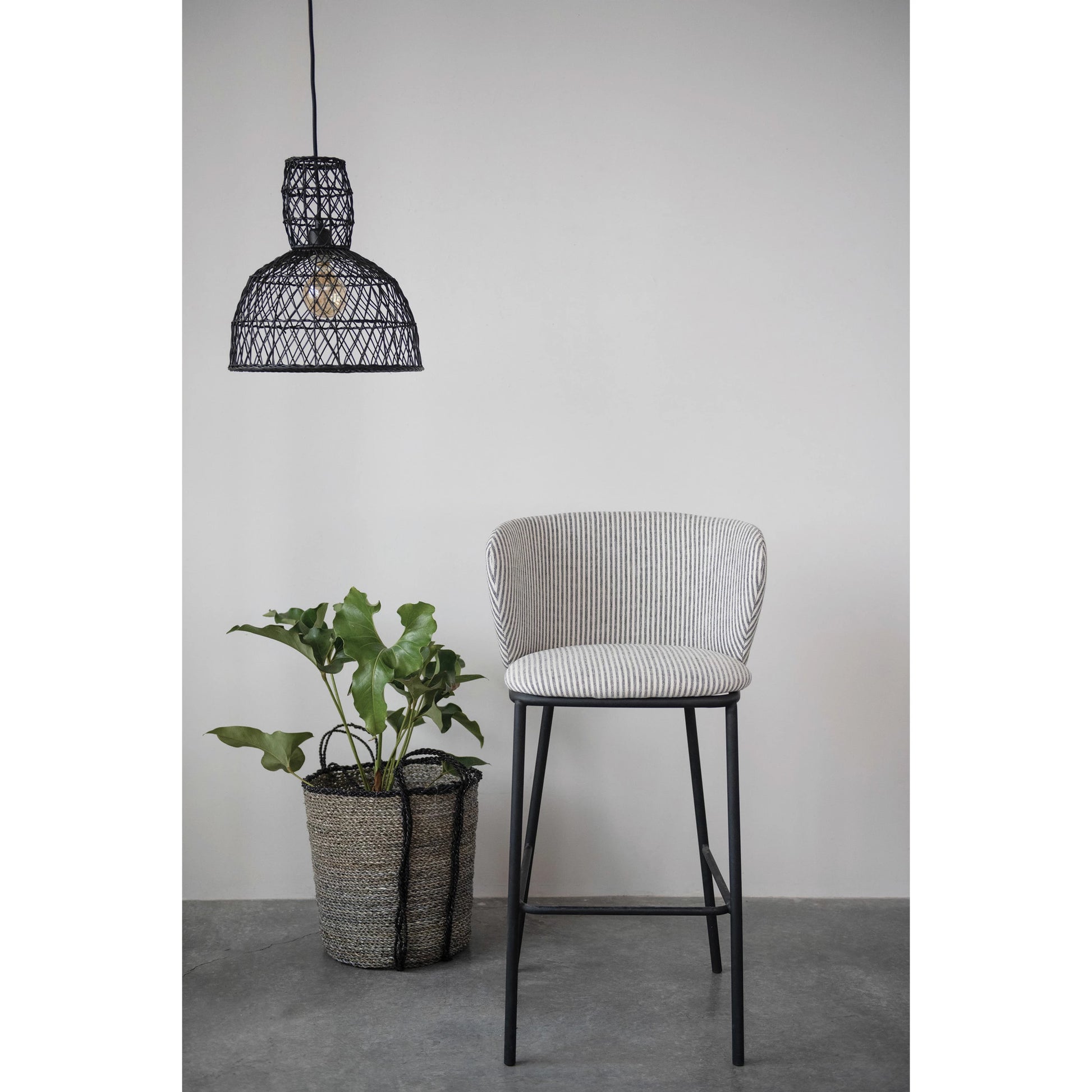 woven rattan and metal pendant lamp hanging above a chair beside a basket with a tall plant inside against a light gray wall
