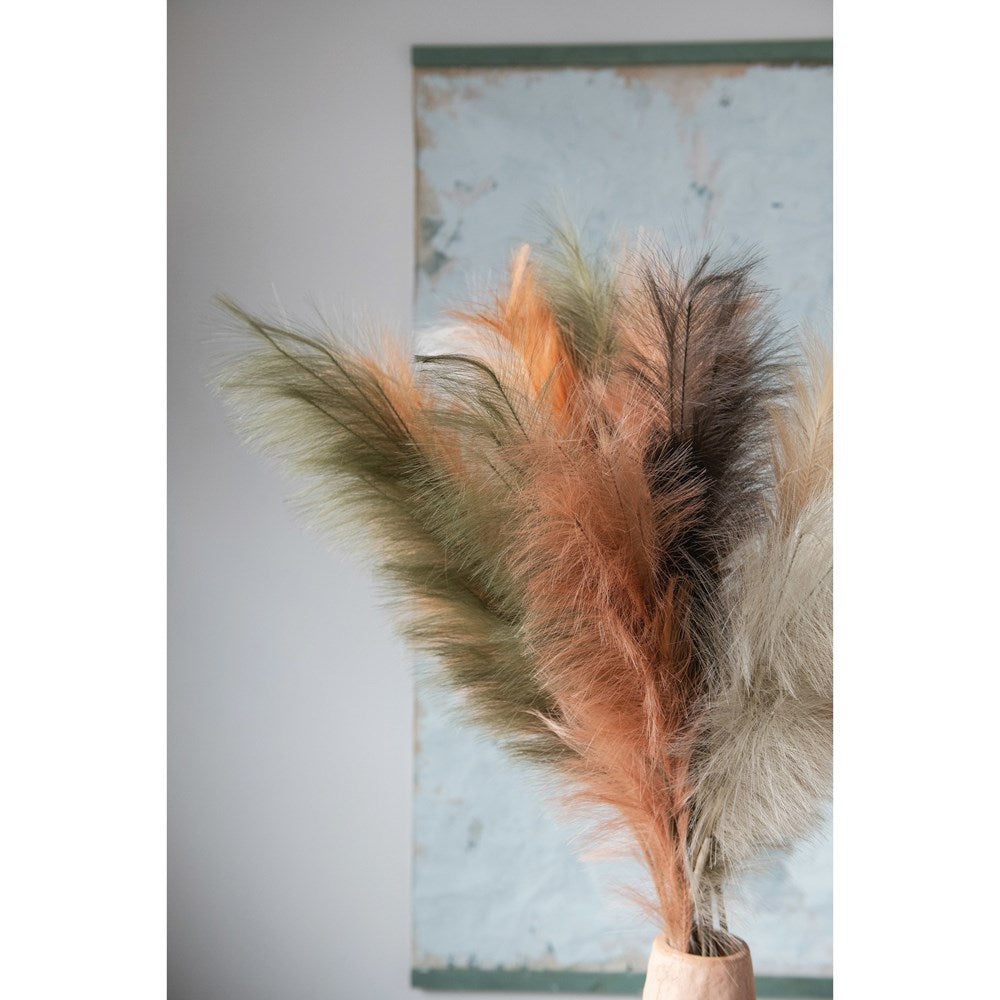 multiple colors of pampas grass plumes displayed in a wood vase against an abstract painting