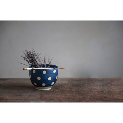 stoneware noodle bowl and chopsticks displayed with small twigs inside on a dark stained table against a gray background