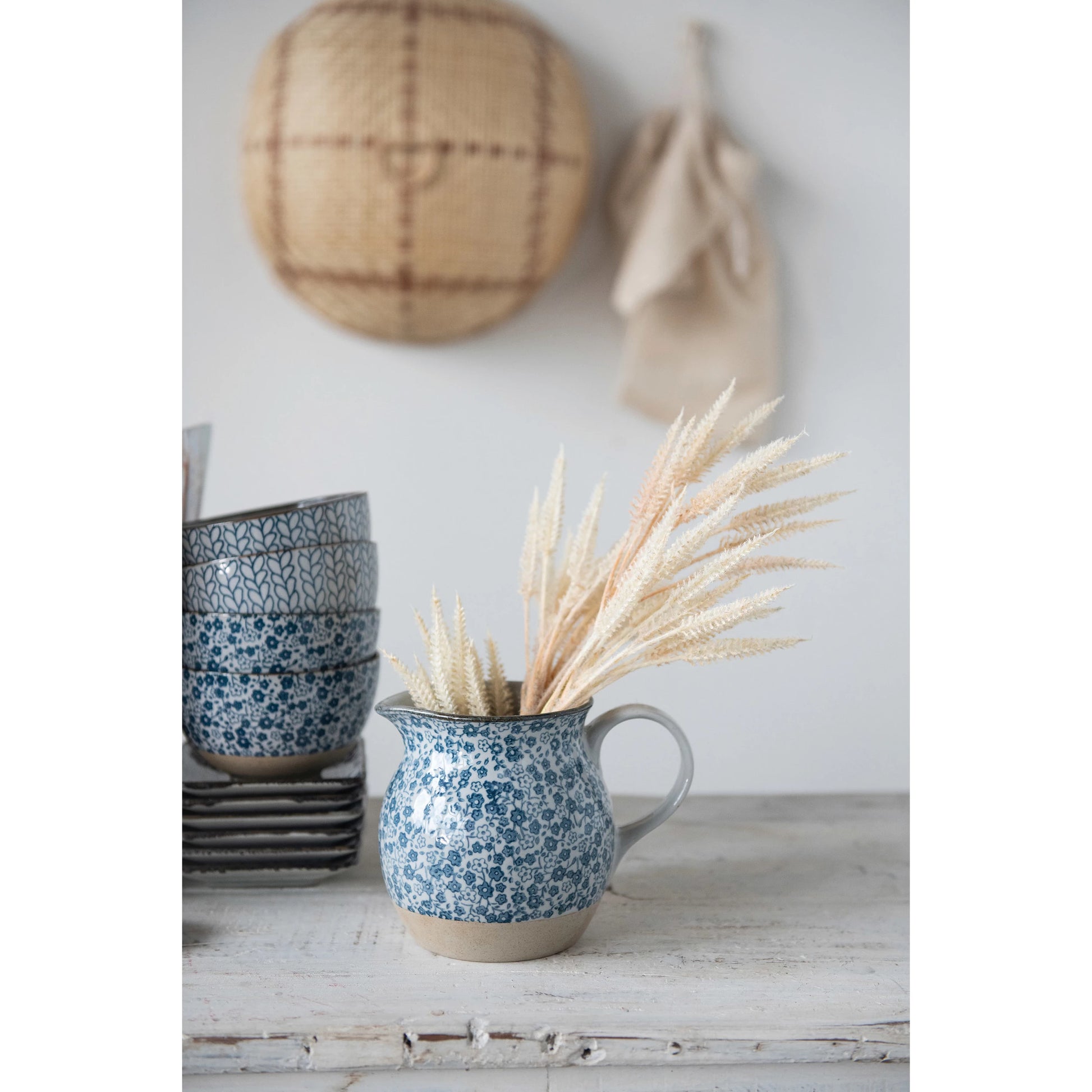 blue floral print stoneware pitcher displayed with wheat stems inside next to a stack of square plates and bowls on a whitewashed wood table with a round basket and towel hanging on the wall in the background