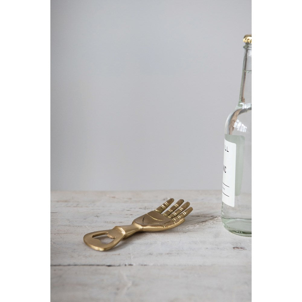 brass hand bottle opener displayed on a white wood table next to a bottle against a gray background