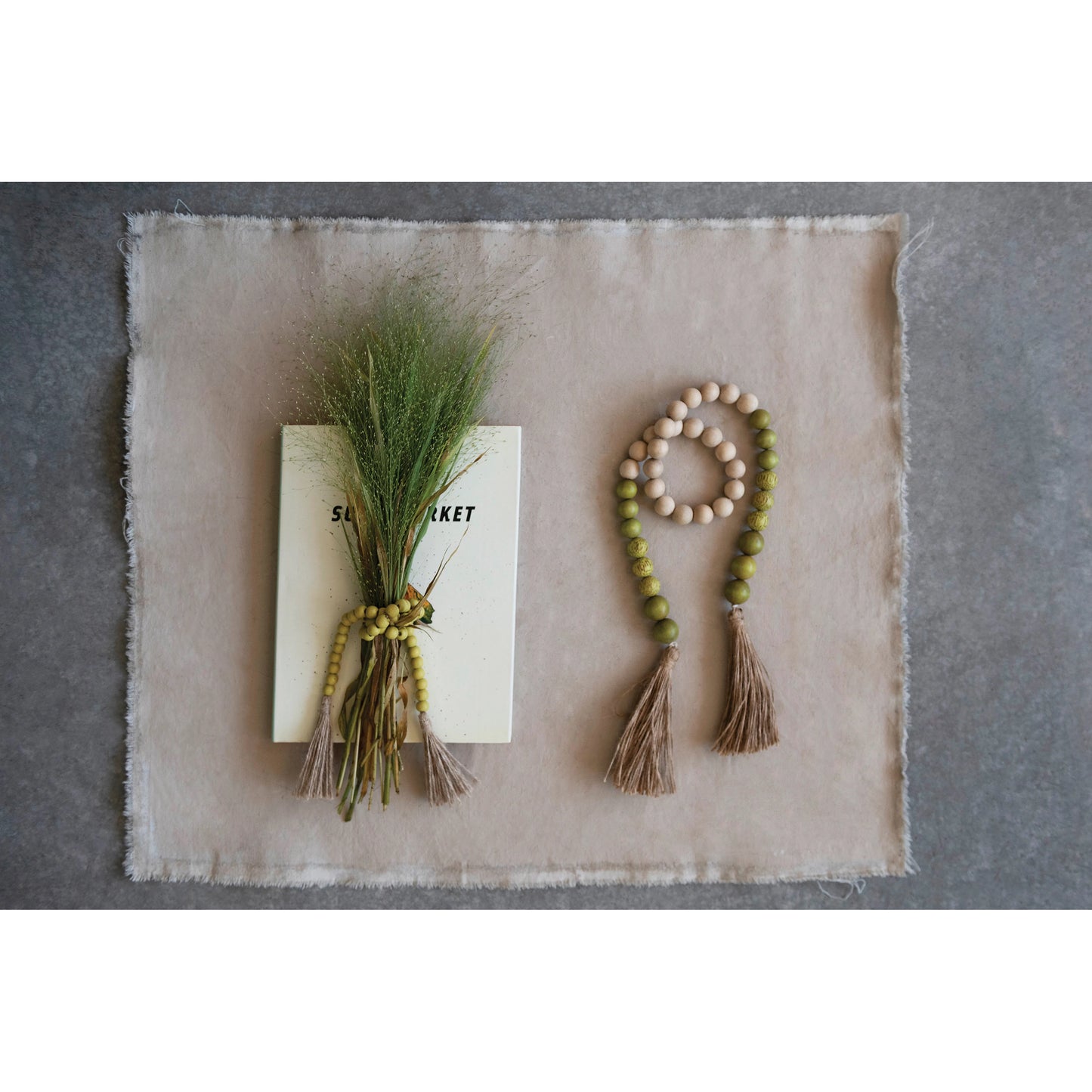 wood and coco shell garland with jute tassel displayed next to a book with a greenery bunch on top sitting on a stone colored square placemat on a gray surface