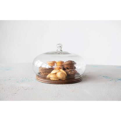 cloche with mango wood base displayed with muffins and cookies inside on a white textured surface