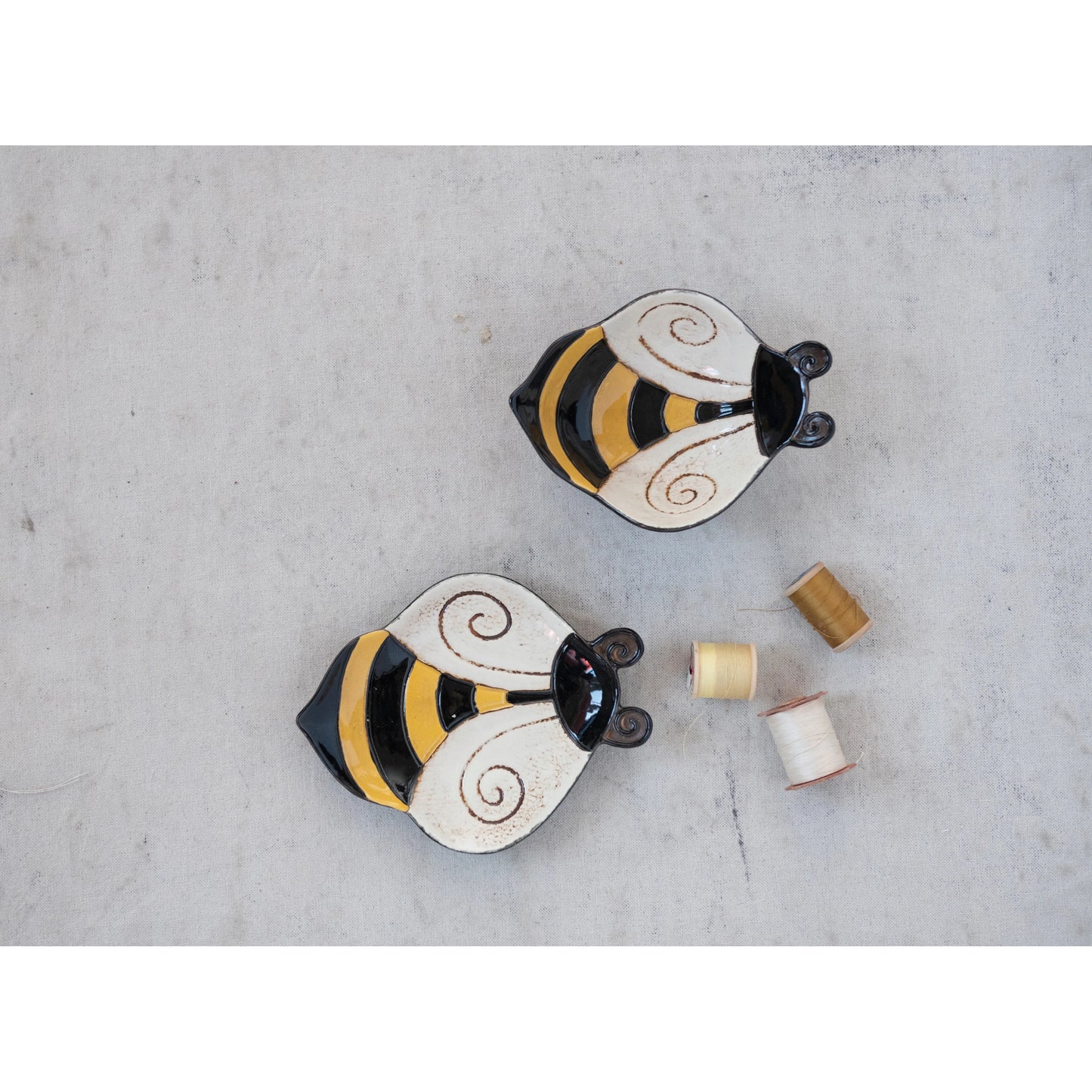 two hand painted stoneware bee bowls displayed next to three spools of thread on a light gray background