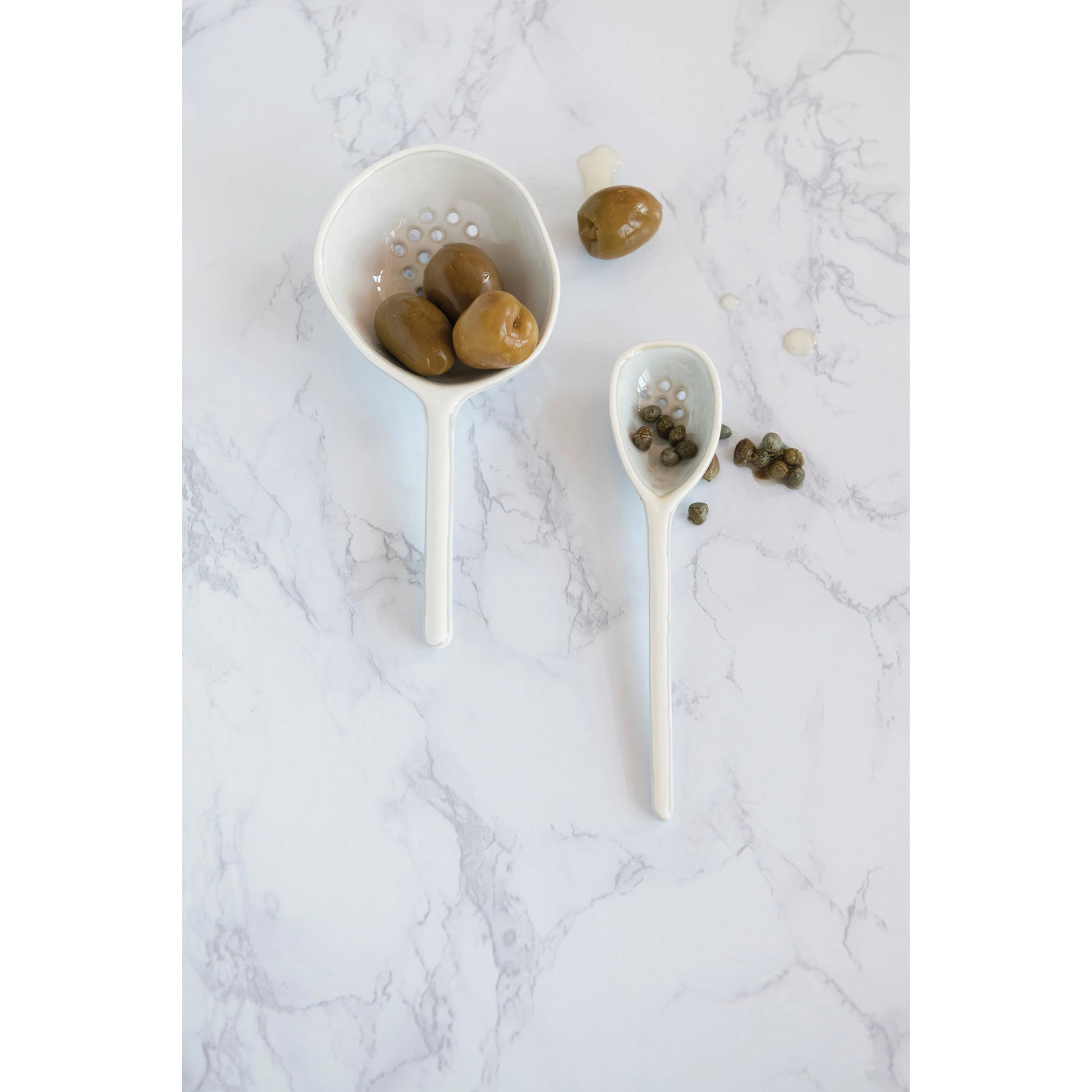 white ceramic straining spoons filled with olives and capers on a grey marble background.