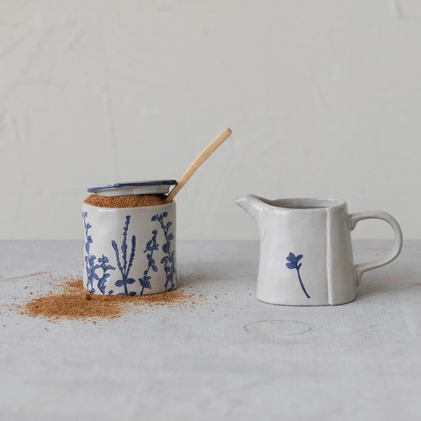 off-white cream and sugar set with blue floral patterns on a wooden table. sugar jar is filled with spice and a wooden spoon.