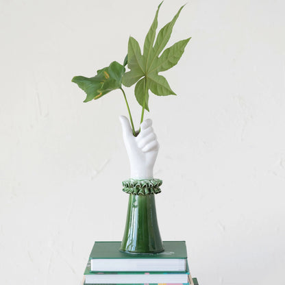 vase shaped like a hand holding 2 large leaves wearing a green ruffle cuffed sleeve set on a stack of books.