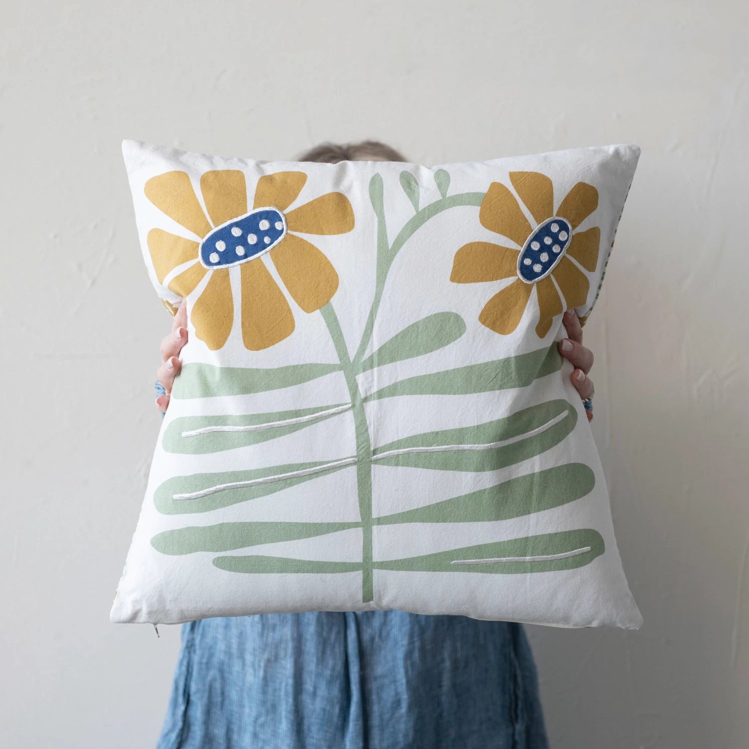 person holding a pillow up in front of their face. pillow is off-white with 2 yellow flowers near the top on green stems with blue centers.