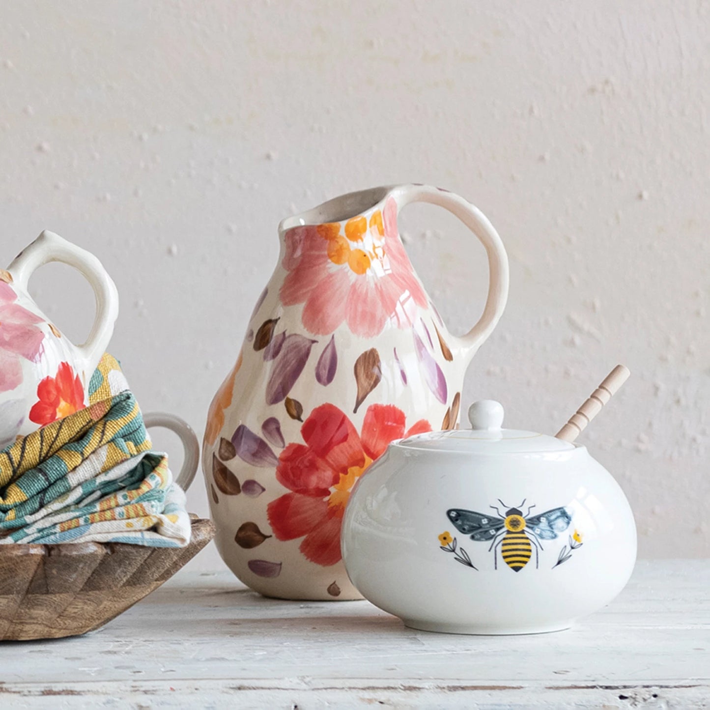 hand painted floral stoneware pitcher displayed next to a bowl filled with folded towels and sugar bowl on a whitewashed wooden table