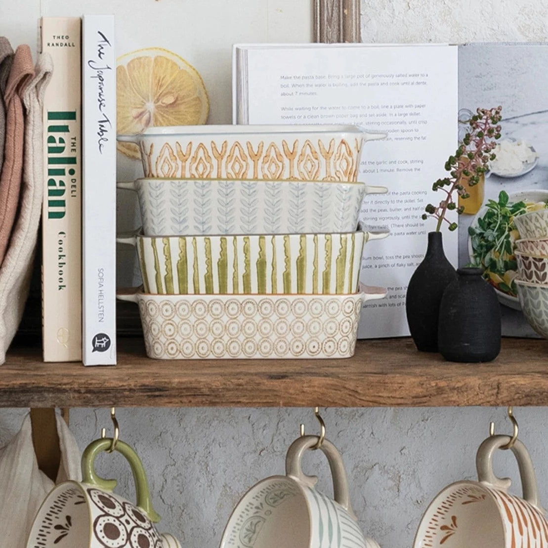 stack of 4 baking dishes on a wooden shelf with books, vases, and mugs on hooks underneath.