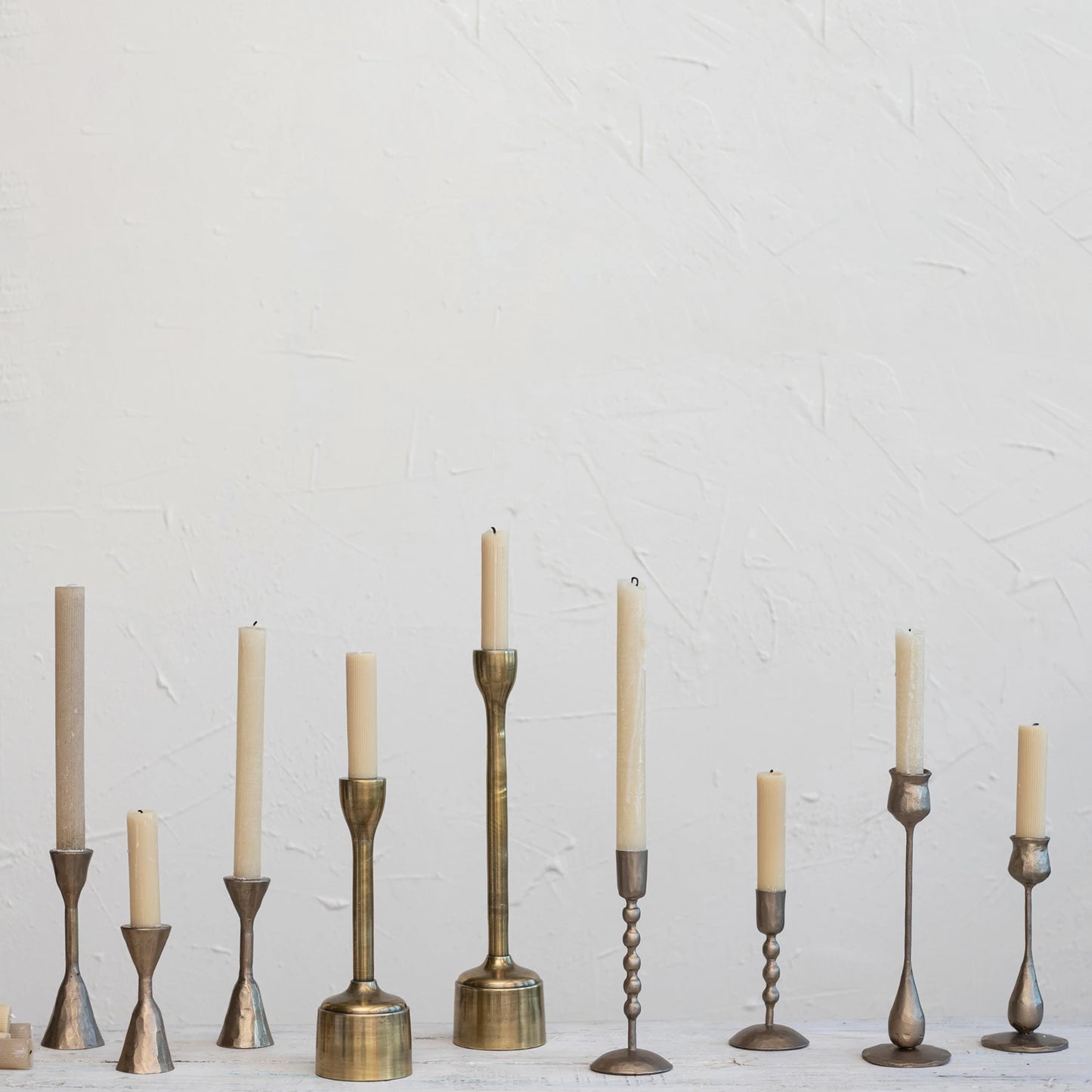 assorted brass candle sticks with cream taper candles in them with an off-white plaster background.