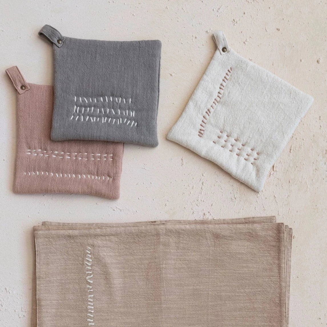 all three colors of hand embroidered cotton slub pot holders displayed next to hand towels on a light tan surface