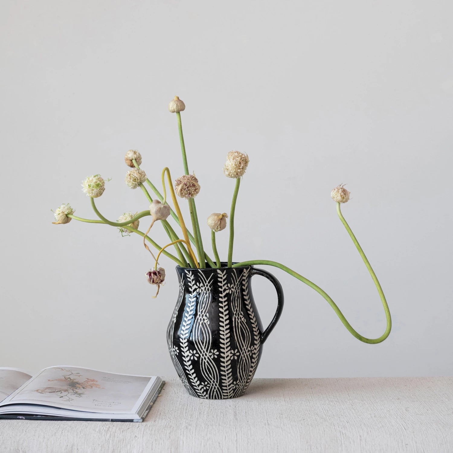 black and cream pitcher filled with onion grasses set on a table next to an open book.