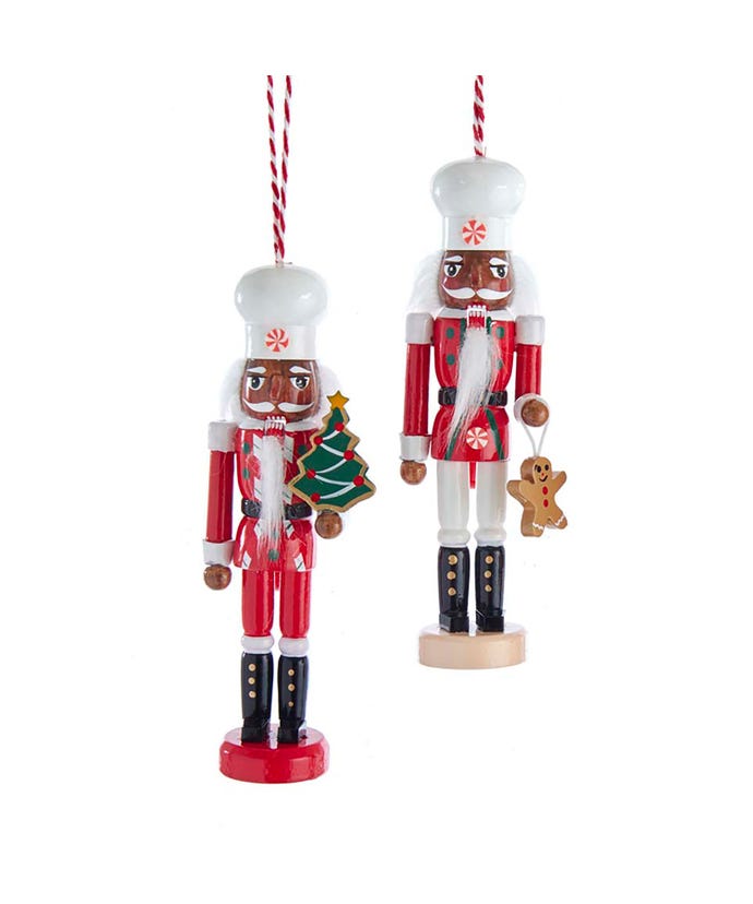 2 wooden nutcracker ornaments with dark skins tones wearing white chef hats and black boots, one has on a red coat and red pants and is holding a tree, the other has a red coat, white pants, and is holding a gingerbread man.