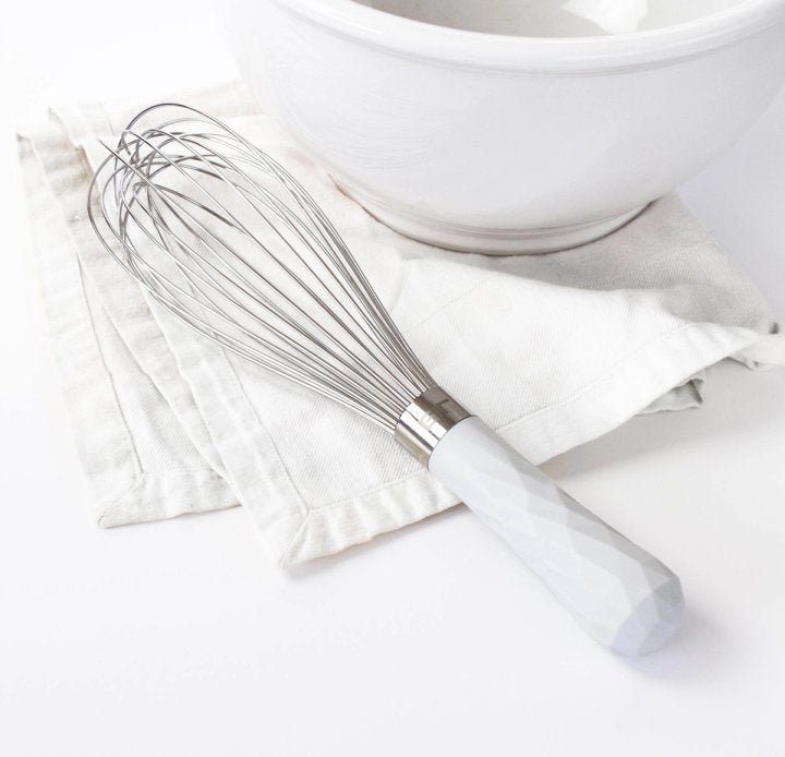 ultimate whisk laying on a white dishtowel next to a mixing bowl on a white countertop.