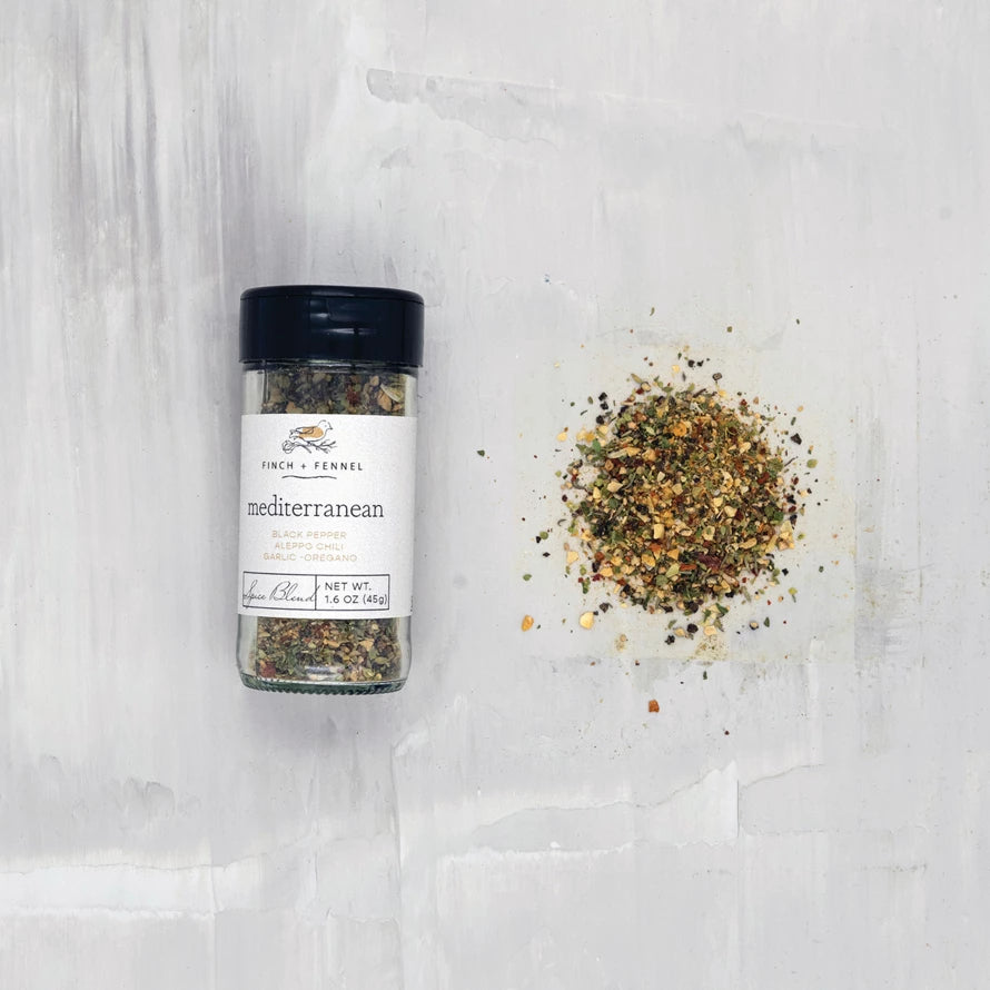 jar of mediterranean spice blend laying down next to a scatter pile of seasoning on a white surface