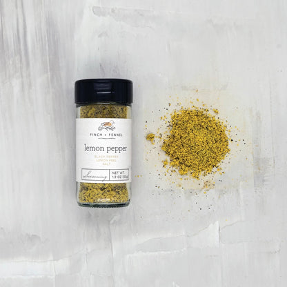 jar of lemon pepper laying down next to a scatter pile of seasoning on a white surface