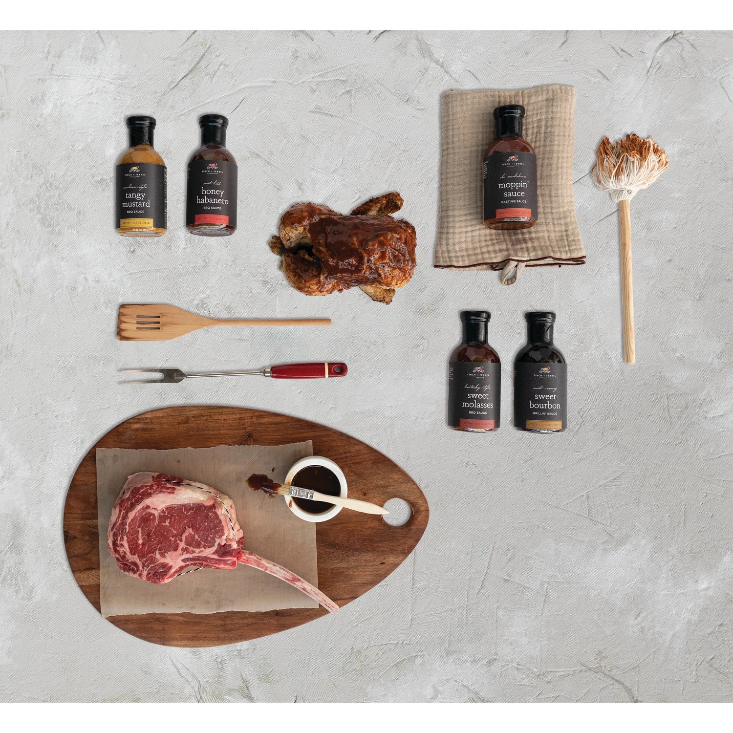 multiple bottles of sauce laying next to a cutting board with a uncooked steak, grilling tools, grill mop and towel on a light gray surface