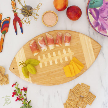 football shaped serving and cutting board displayed with snack foods and utensils on a mostly white background