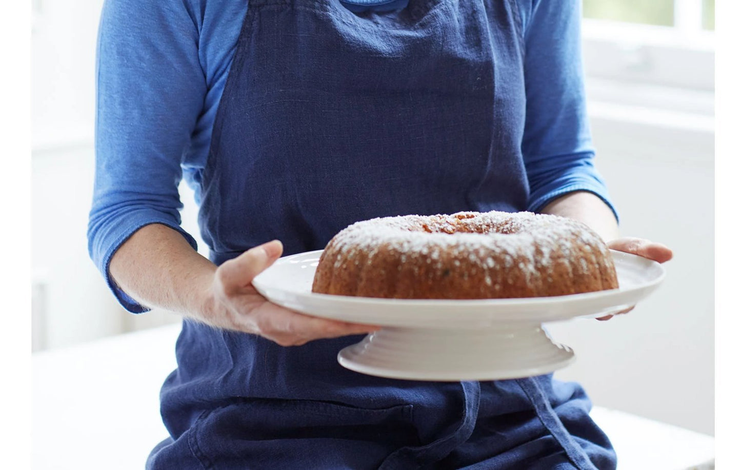 person wearing blue apron holding cake plate with cake on it.