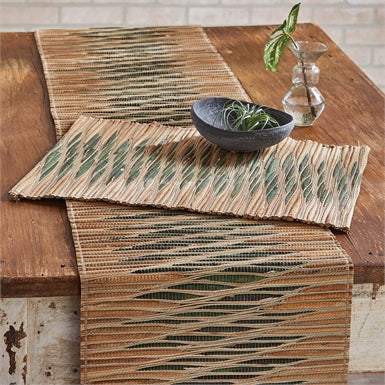 table runner hanging over the edge of a wooden table with a placemat, bowl and glass vase on it.