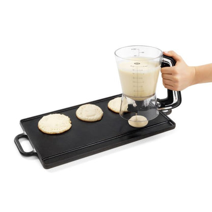 hand holding batter dispenser over gridle dispensing batter for pancakes.