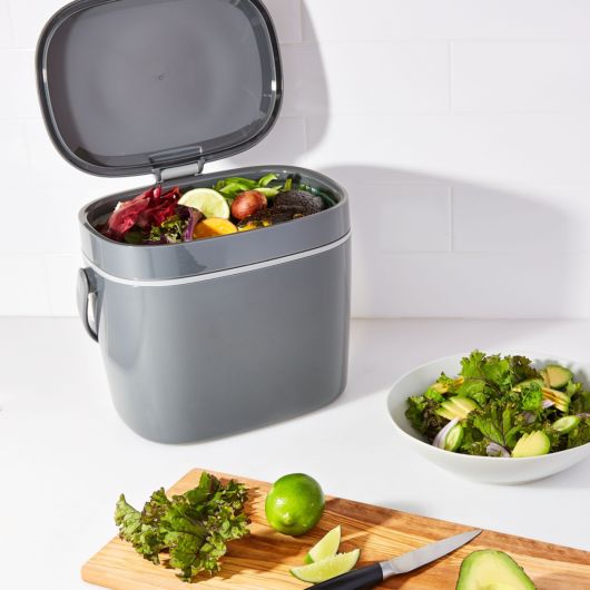 compost bucket open on counter with food scraps in it and salad ingredients in a bowl and on a cutting board.