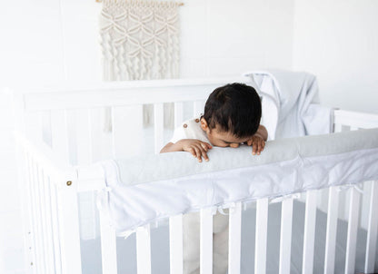 a baby boy standing and chewing on the grey crib chomper in a child's room