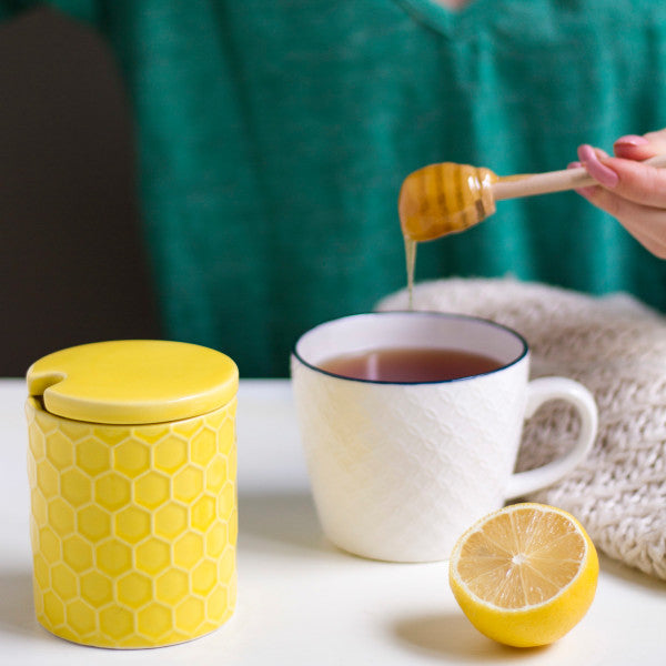 yellow honeycomb pot set next to a a white mug of hot beverage with a hand drizzling honey into it and a sliced lemon.