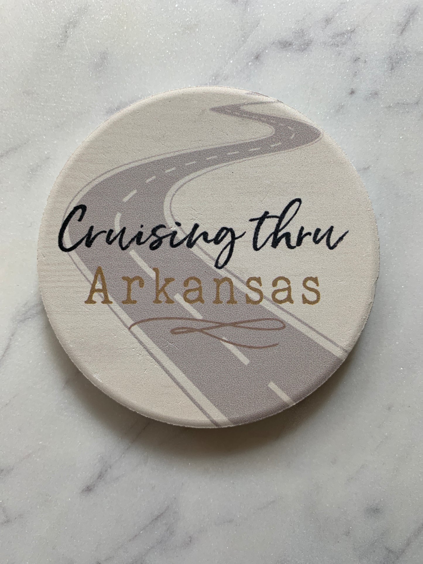 cruising thru arkansas car coaster is beige with a gray road across it and text in black and gold displayed on a gray and white marble surface