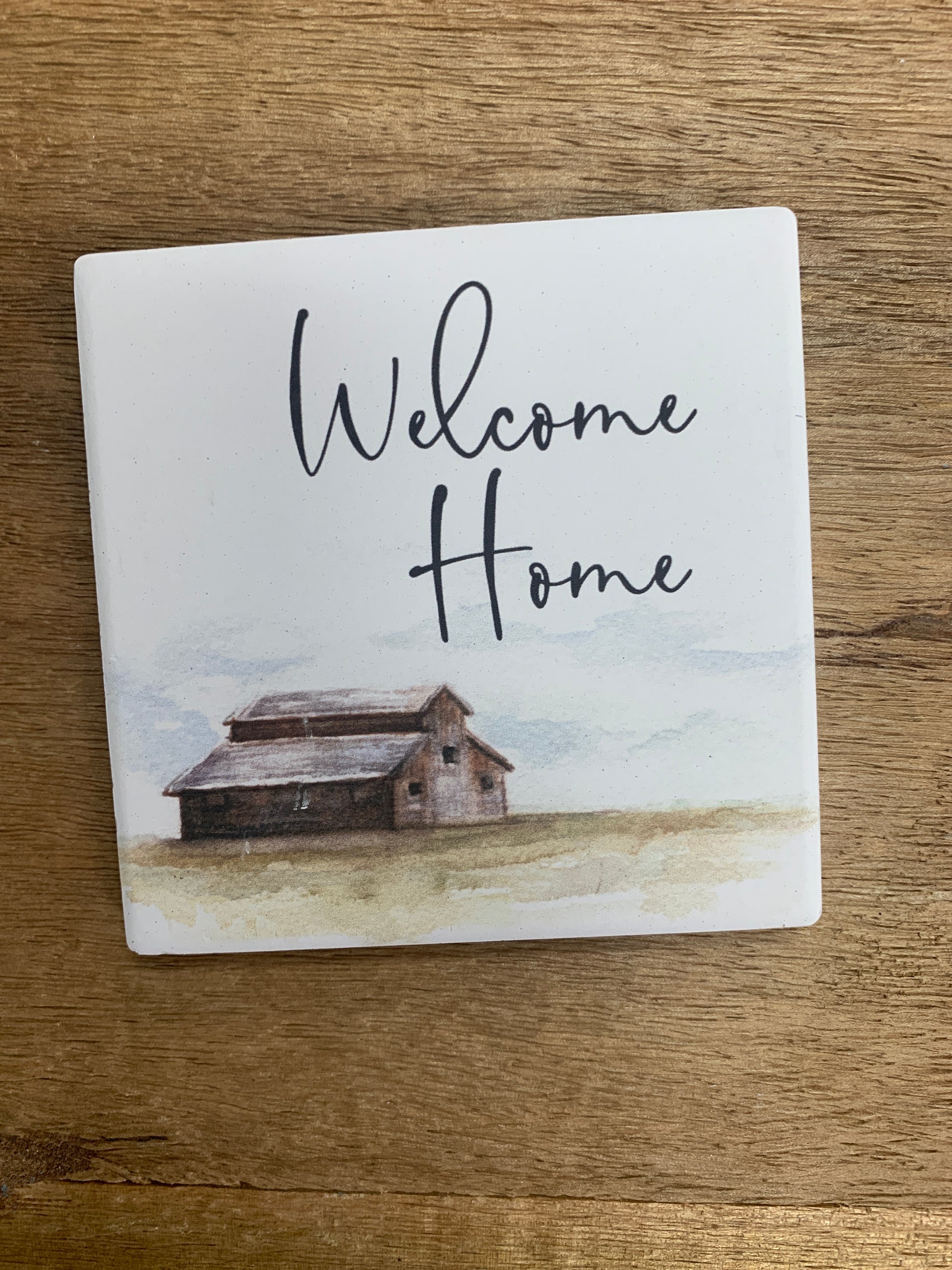 welcome home coaster is white with a barn in the distance with black text displayed on a wooden surface