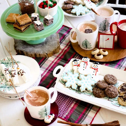 an arrangement of holiday platters, plates, and mugs on a tabletop with a plaid runner, cookies, and hot cocoa.