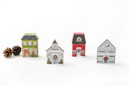 3 printed wood block houses and 1 church decorated with images of wreathes, greenery, snow, and lights set on a table with pinecones and artificial snow.