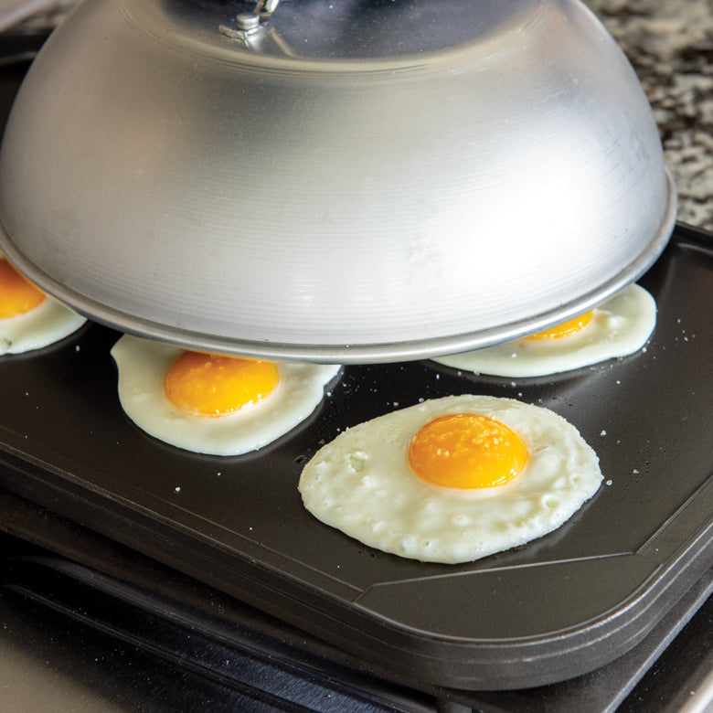 domed lid over griddle with fried eggs on it.