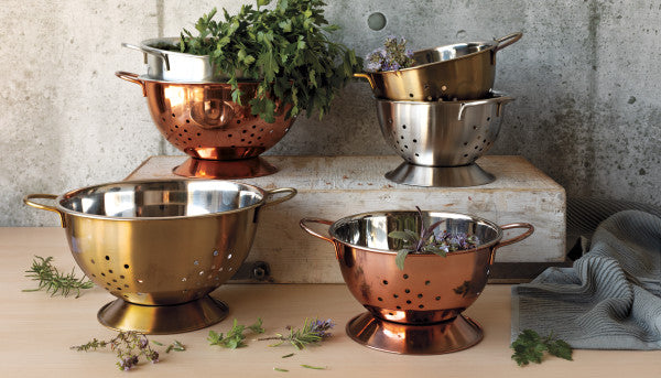 6 colanders arranged with herbs and dishtowel on countertop.