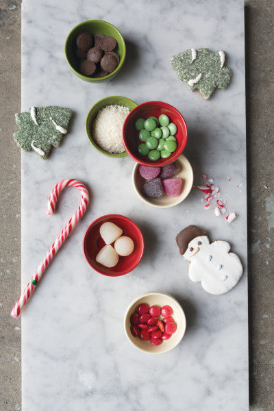 top view of all 6 holiday pinch bowls filled with different candies, next to a snowman and tree cookies, and a candy cane on a white and marble gray surface