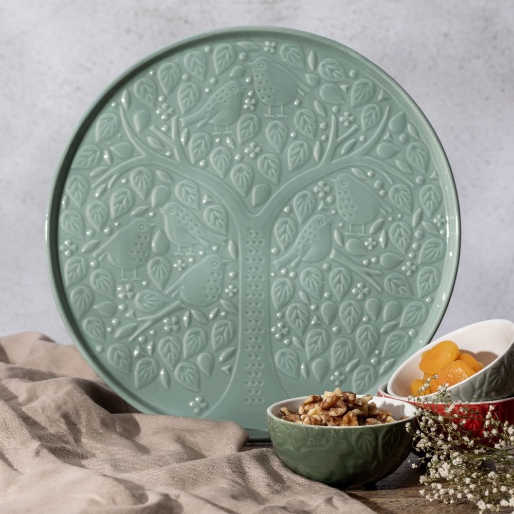 green platter with woodland scene embossed on it leaning on a countertop with small bowls of fruit and nuts and a towel set next to it.