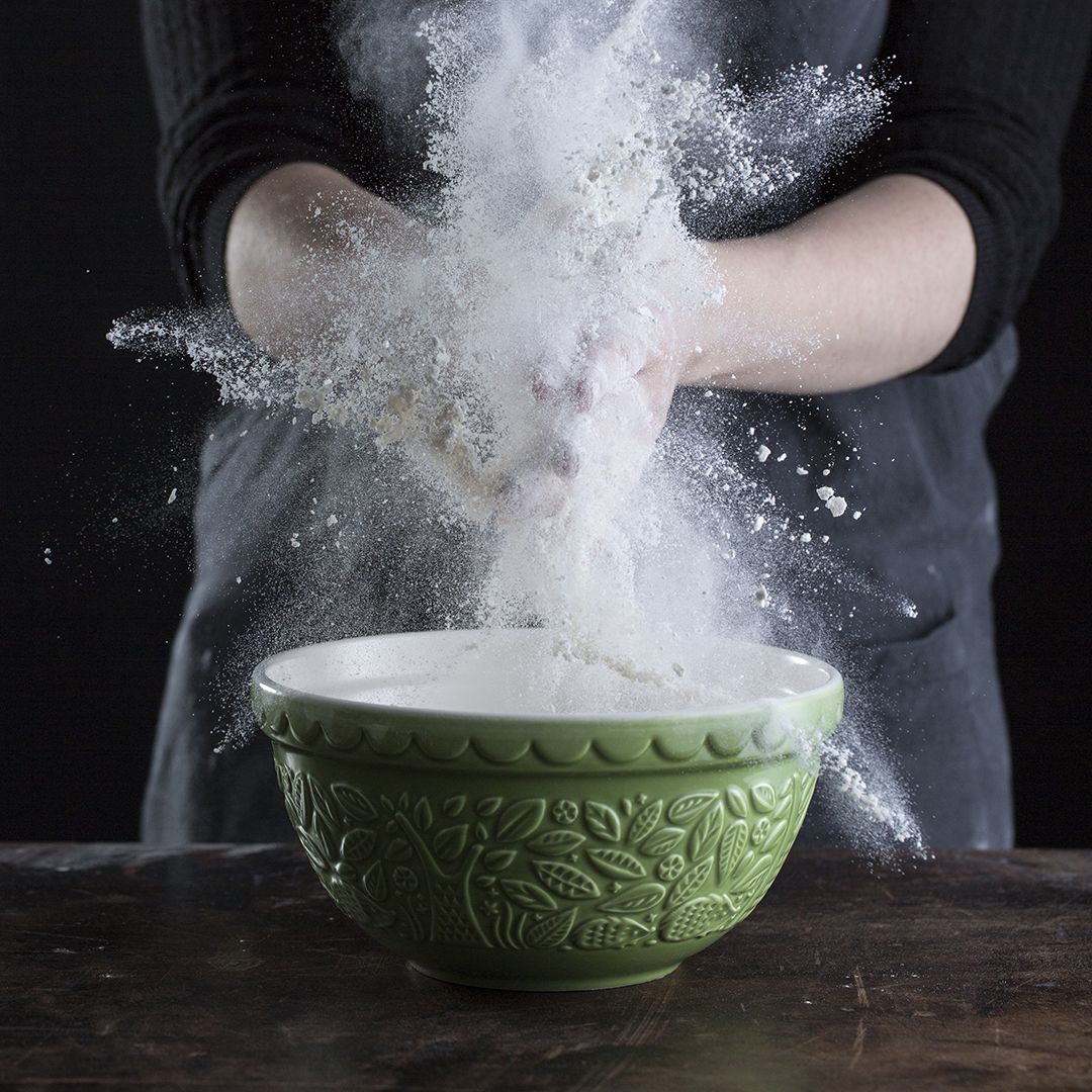 hands adding flour to mixing bowl on wooden countertop.