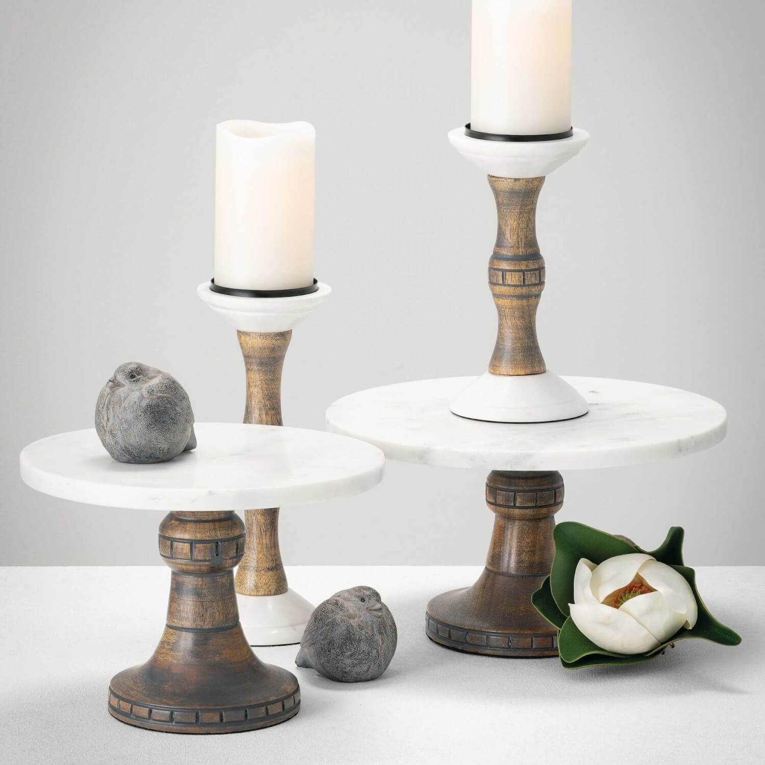 2 marble-topped pedestals with wooden bases styled with bird decor, faux flowers, and candles on similiar candle pedestals.