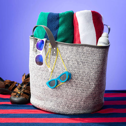 dove gray woven textile basket displayed with rolled beach towels, sunscreen, and sunglasses next to a pair of sandals on a red and blue striped beech towel