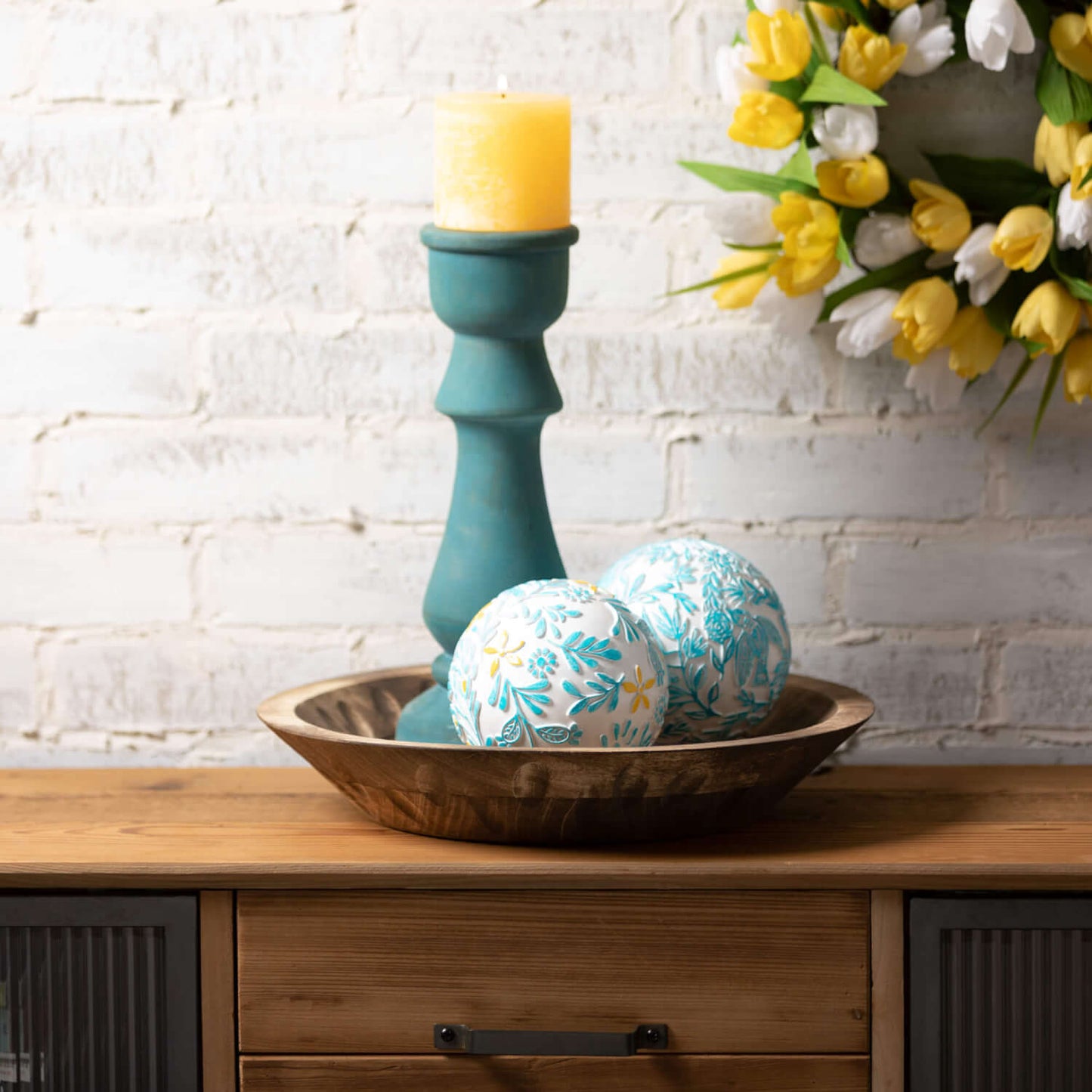candle stick with yellow candle on it set in a wooden bowl with 2 decorative orbs.