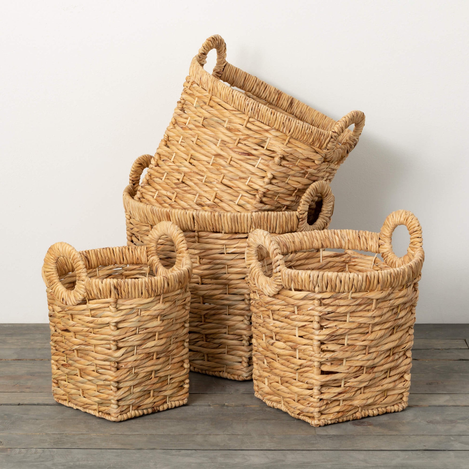 4 sizes of woven seagrass baskets with round handles arranged on a wood floor with white wall in background.