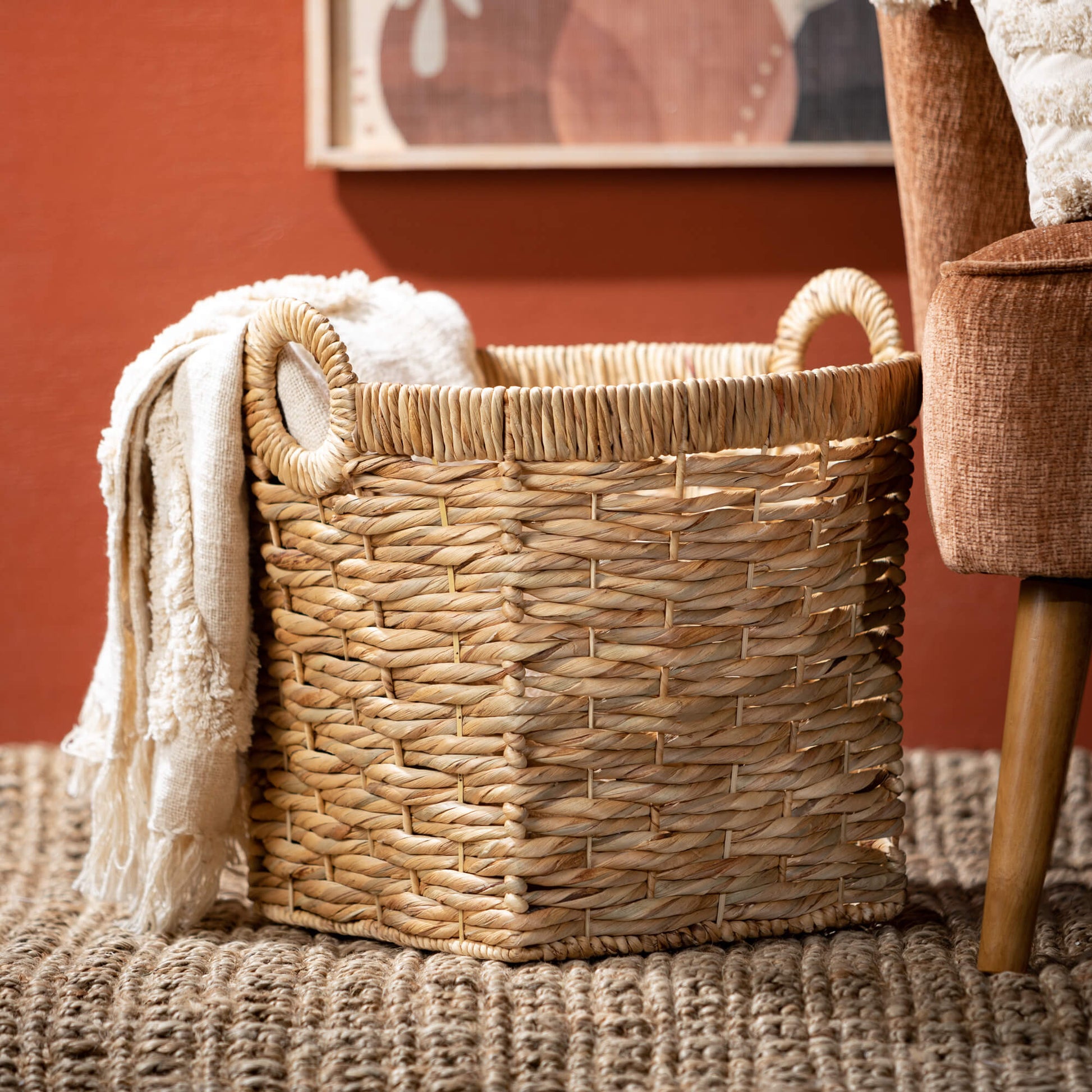 basket on the floor with a blanket draped out of it set next to a chair.